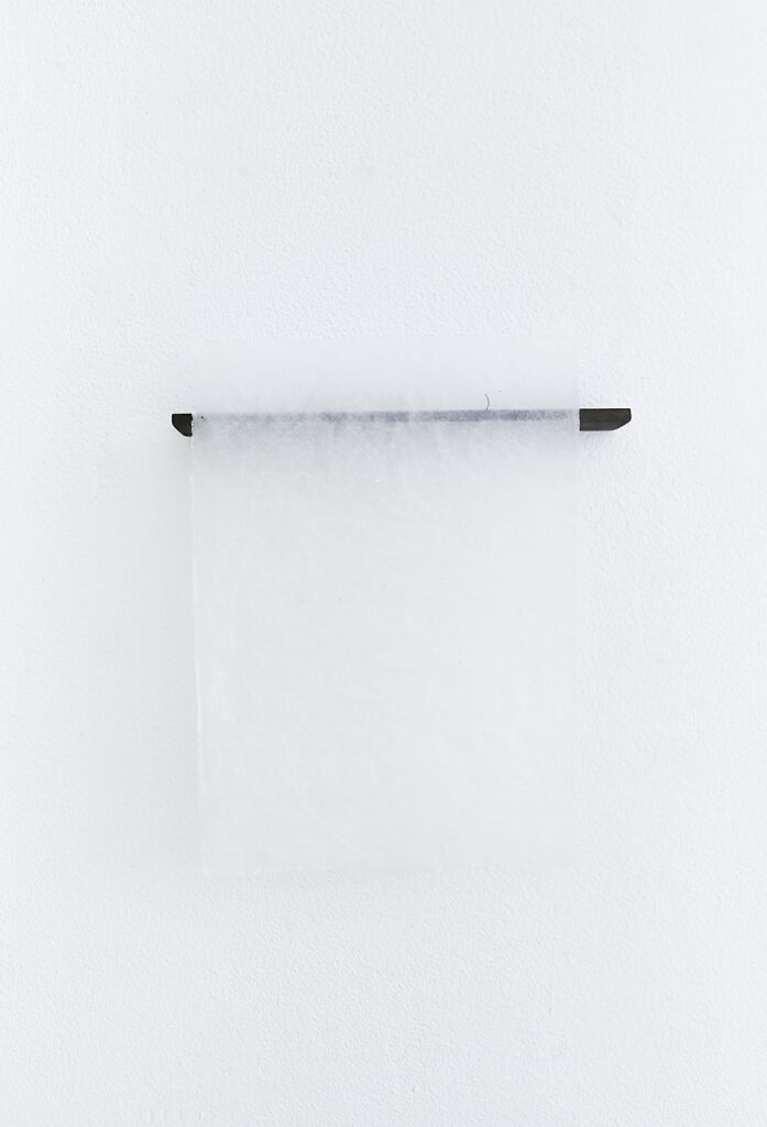 a black piece off wood with a transparent handmade paper over it is haning off a white wall